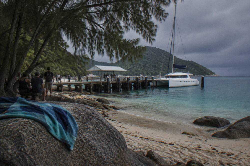A white catamaran ferry moored at a tropical island jetty with passengers waiting in the shade