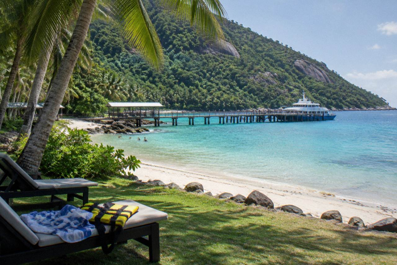 A sheltered tropical bay with a jetty, turquoise shallows and rainforest covered slopes rising behind the beach
