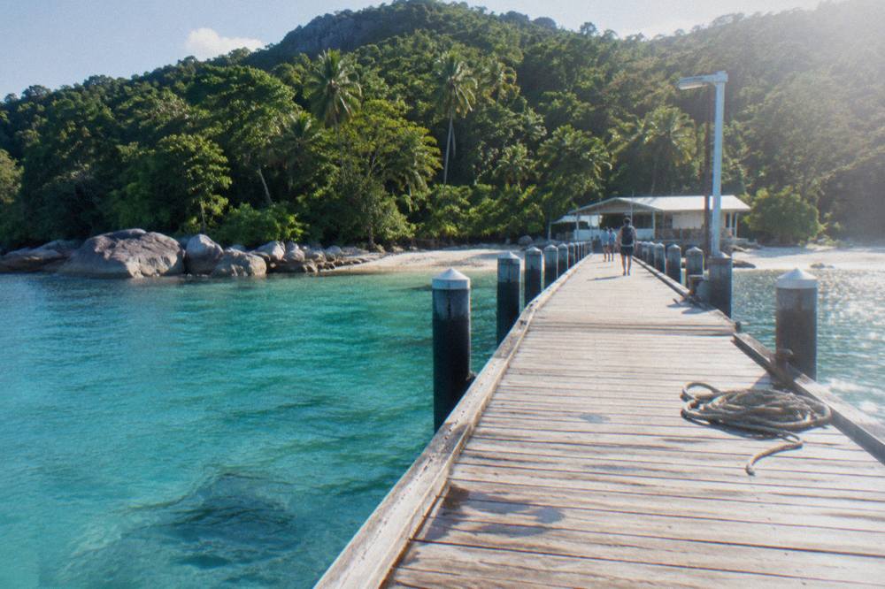 A wooden jetty stretching over clear turquoise water towards a forested island