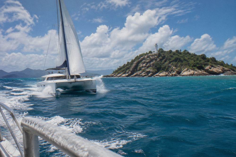 A white catamaran cruising past a green forested island on calm blue sea