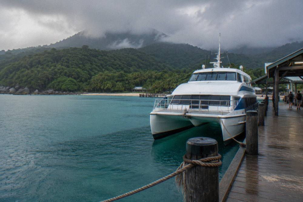 Fast catamaran ferry moored at a tropical island jetty with rainforest covered hills behind