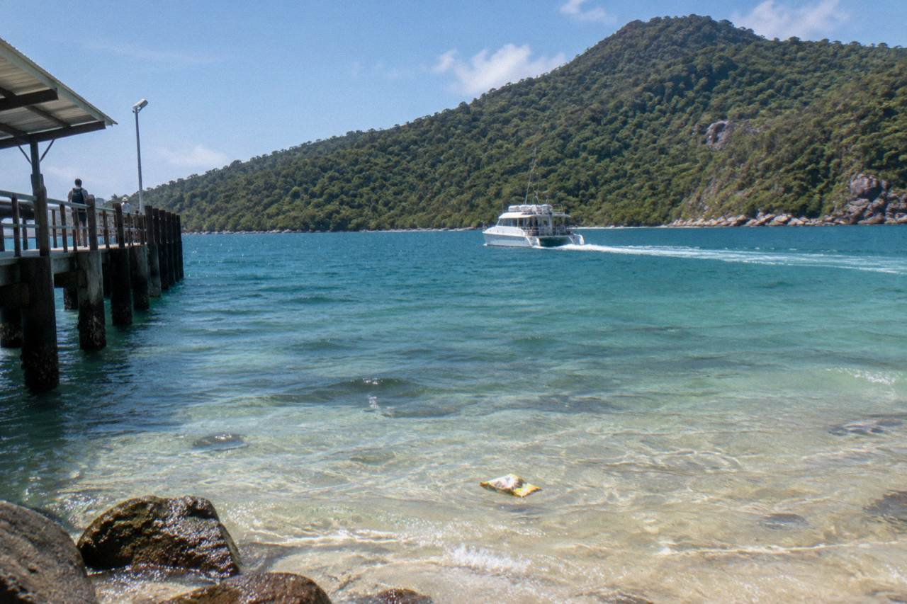 A catamaran ferry crossing calm blue water toward a green tropical island fringed by coral beach