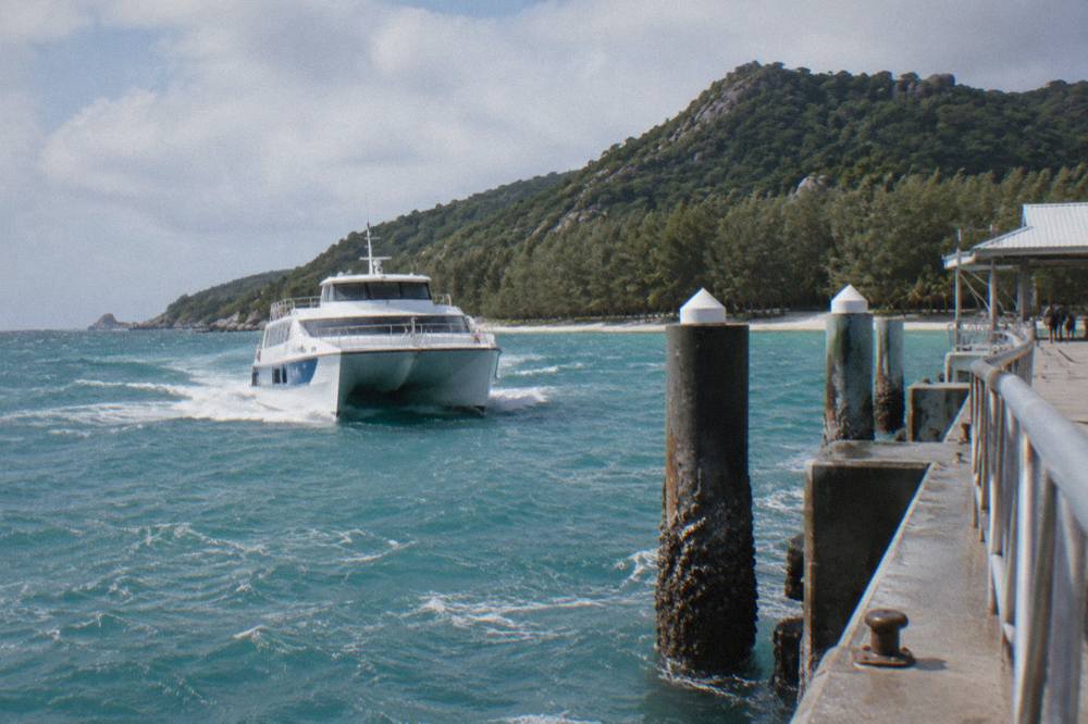 A fast catamaran ferry approaching a tropical island jetty across calm blue sea