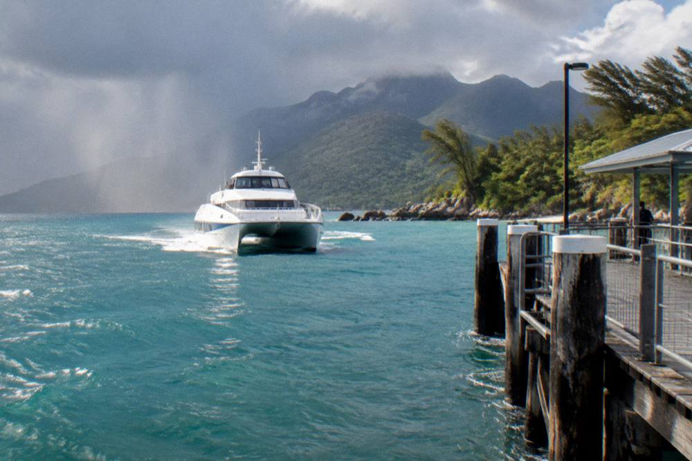 A fast catamaran ferry approaching a tropical island jetty across calm blue sea