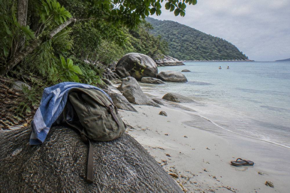Day pack and towel on a wooden bench beside a coral beach with rainforest beyond