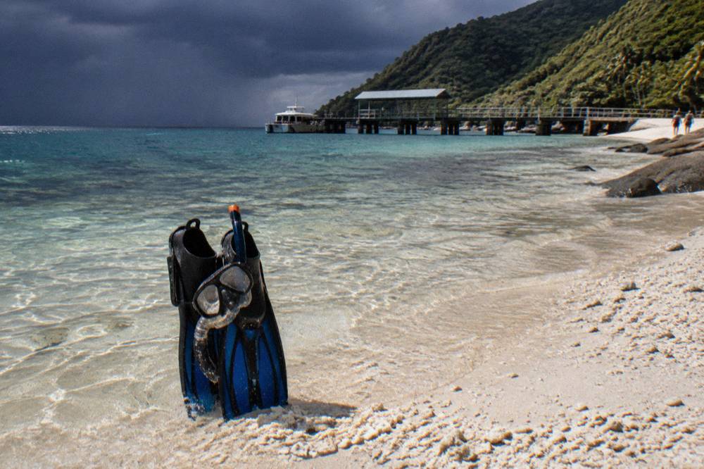 Snorkel mask and fins resting on white coral sand beside clear shallow water